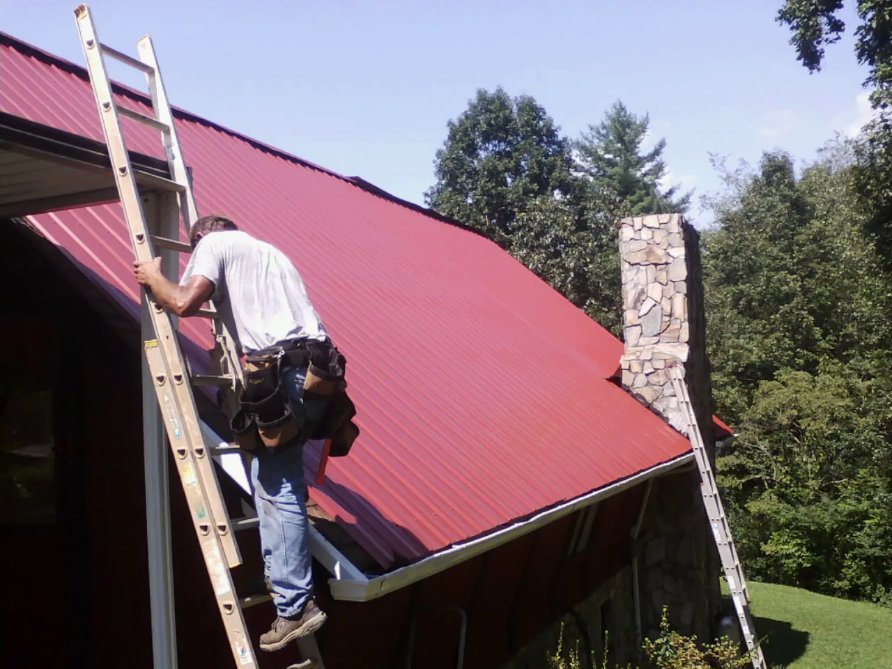 Roofer on a ladder beside a red metal roof and stone chimney — Soffit & Fascia Repair by Roofing Brothers in Owasso
